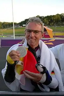 Man with glasses draped in a German flag holding up a silver medal.