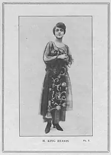 Full length photograph of a standing woman wearing an ankle length dress in which the top layer is sheer and has art deco motifs as embellishment