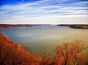 View of a very wide river lined with bare trees in reddish light under a blue sky streaked with clouds