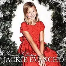 Evancho, with her straight blond hair falling on her shoulders, sitting in a large Christmas wreath, facing the camera, and wearing a red party-dress.