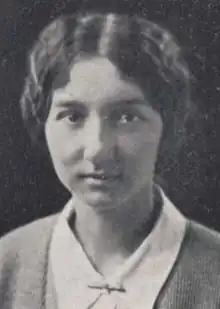 A young woman with dark wavy hair and dark eyes, wearing a collared shirt, a cardigan, and a bar-shaped pin