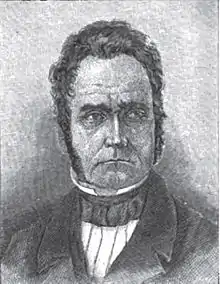 A man with curly, black hair and sideburns wearing a black jacket and white shirt