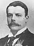 A black-and-white portrait of a white gentleman in a dark suit, a carnation prominent in his buttonhole. He has a square jawline and a handlebar moustache.