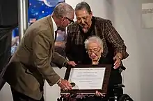 Johnson seated in a chair holding a NASA Group Achievement Award flanked by a man and a woman