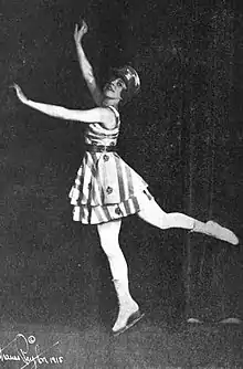 A young white woman in ice skates and a striped skating dress, arms and legs stretched in a dancerly pose