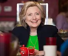 Hillary Clinton dressed in a black suit and a green shirt, sitting in a café. She is smiling, and a red teacup is situated in front of her. The foreground is distorted due to the presence of various small objects.