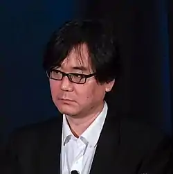 A middle-aged Japanese man with glasses in front of a microphone