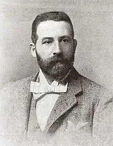 head and shoulders photo of white man of middle age, with neat dark hair and full-set moustache and beard neatly trimmed