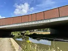  A brick and concrete road bridge spanning a canal with a dirt path on the left side, occupied by a cyclist in the distance