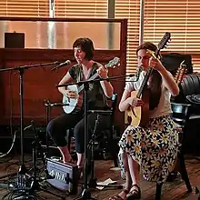 Two female musicians, one with a banjo and one with a guitar sit in front of microphones