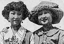 A black-and-white newspaper photograph of two young Asian women, side by side, photographed outdoors. The woman on the left is not smiling or wearing a hat; the woman on the right is smiling and wearing a hat.