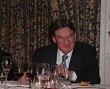 A formally dressed man sits at a table with several wine glasses on it