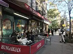 View of cafe from sidewalk with red awning, outdoor seating, and colorful murals on exterior walls.