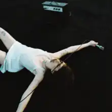 Jilian Medford wearing a white dress, lying on her back on a black floor with a microphone in her hand