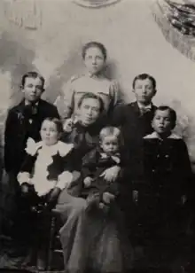 A family portrait of Ida Hunt Udall with her children. All are dressed formally. Udall wears a fine, black dress. In her lap she holds her youngest sons, Gilbert and Don Taylor. Her other children stand behind and around her, roughly in a semicircle. Pauline Udall, the oldest of the children, wears a dress whose lighter color contrasts with her mother's.