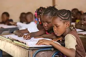 Two young girls sit close together at their desk, and look attentively at their workbook.