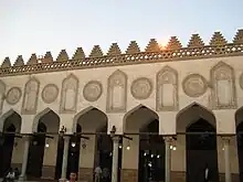 Five keel shaped arches (and part of a sixth) are visible. The arches are supported by cylindrical columns. Above each arch is a large circular inscribed stucco decoration, and above each column is a large inscribed stucco decoration that mirrors the shape of arch and columns. Behind the row of columns is a walkway, and then a wall with entrances the shape and size of the arches and columns.