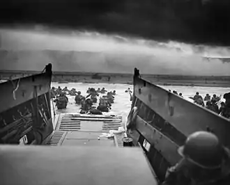 A black and white image of troops leaving an amphibious troop transport. They are walking through the water to a beach.