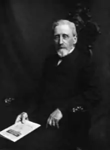 Man in dark suit, seated in an armchair, holding a pamphlet.