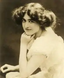 A young white woman with dark hair in a bouffant updo, seated with one hand on chin