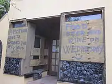 Entrance of a bar with its decorated glass windows boarded with two wooden boards. The first board on the left reads, "Another quality board job by Cirós" and the second board reads, "Food beer and wine on Wednesday" with a smiley emoticon