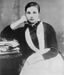 Portrait of a girl sitting in a chair leaning her right elbow on a small table stacked with books, who is wearing a dark dress and a white apron