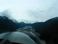 To the right and left are mountains, between which is a deep valley with numerous meandering and intersecting segments of the Iskut River proceeding into the distance. Beyond it are snow-capped mountains.