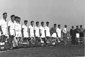 A black-and-white photograph of a football team lining up before a match. Eleven men in old-fashioned association football attire stand in a line: ten wear white shirts, white shorts and black socks, and the other wears a dark shirt. Beside the players stand two suited men, and beside them stands another player in different colours, his dark shirt marked "CCCP".