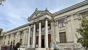 The façade of a masonry building, with four Greek adorning its entrance, under a clear blue sky