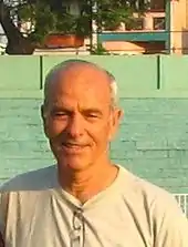 An upper-body photograph of a white man with balding grey hair, smiling at the camera while wearing a grey shirt.