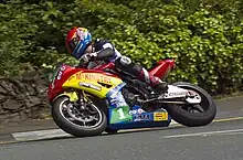 Rider of a multi coloured racing motorcycle against a background of heavy foliage exiting Governor's Bridge Dip, a right turn on the Isle of Man roads in the capital town, Douglas, promoted to city status since 2023