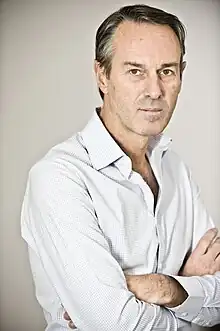 Color portrait photo of a white man wearing a long-sleeved shirt, standing with his arms folded.