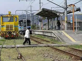 A passenger uses the level crossing to reach the island platform. On the left, the passing siding is being used by a track maintenance vehicle.