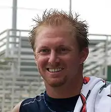 A smiling young man with short, spiked red hair and a towel laid over his left shoulder