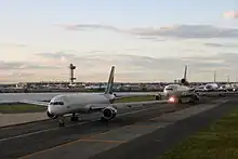 Five jumbo airplanes wait in a line on a runway next to a small body of water. Behind them in the distance is the airport and control tower.