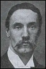 A head and shoulders view of a dark-haired man with a moustache, wearing a suit jacket and wing collared shirt