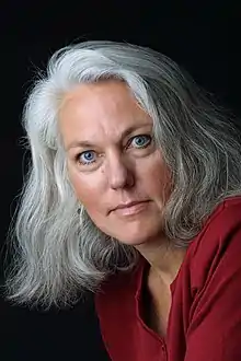 A white woman with blue eyes and silver hair looks into the camera; she is wearing a red top