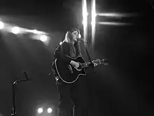 Woman performing onstage, playing guitar against a black background