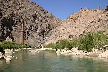 A tall minaret in a river valley. At the top of the nearby mountains there are other, smaller structures.