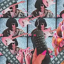 A woman's hand with pink fingernails holds a black television remote in front of duplicates of electric guitars and a picture of James Ferraro with sunglasses and lipstick. On the far right is a woman's hair.