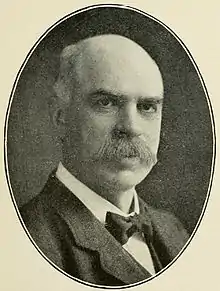 A black and white photo of a bald white man with a bushy mustache and a bowtie gazing sternly at the camera.