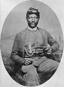 A black man sitting in a chair, wearing a forage cap and a suit coat over a vest and holding a revolver in his left hand. On the vest is a watch fob chain and a circular medal hanging from a ribbon.