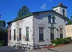 A small brick building with white paint flaking off in spots, green trim, a gently sloping gabled roof and low tower behind the right corner.