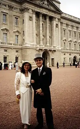 A colour photograph of Jason Tsai, alongside his wife Ruth Tsai.  The couple are dressed in formal wear: Jason is wearing a black morning suit with a grey top hat and matching grey necktie and pocket handkerchief; his wife Ruth is wearing a full-length white dress, matching white jacket and white hat.  The couple are standing in the red-gravelled courtyard of Buckingham Palace - the central London official residence of Queen Elizabeth II, the reigning monarch of the United Kingdom of Great Britain and Northern Ireland.  In the background are other unidentifiable people in similar attire.