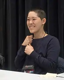 An Asian woman with buzzed black hair and a wide smile wearing a black shirt sits at a table before a black drape. She wears a lanyard and clutches an object, possibly a marker, in front of her chest with both hands.