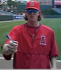 A white man wearing a red baseball cap and warmup, both with a red "A" topped by a halo. The man is holding out his right hand, which is holding a pen, and wearing sunglasses.