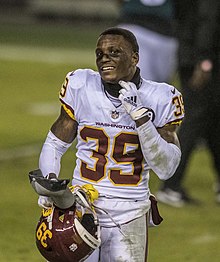 A man in a white football jersey on a green football field looks toward the camera while removing his helmet