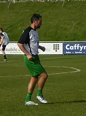 A dark-haired white man wearing sports kit exercising on a sports pitch with advertising boards and a grass bank in the background.