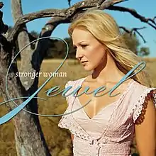 An image of a blonde woman wearing a tan blouse and standing close to a bare tree.