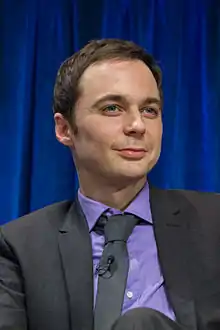 A brunette man wearing a purple dress shirt, black tie, and black jacket stares off into the distance with a slight smirk